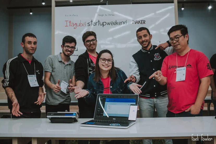 Team of six people smiling and pointing at a laptop with their system open, in front of the Techstars Startup Weekend Maker Itajubá backdrop